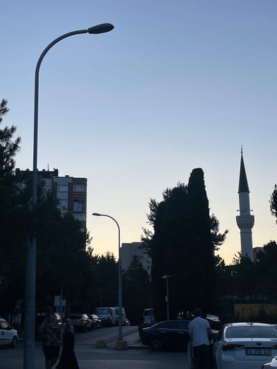 A street scene in Başakşehir, Turkey, features a tall lamppost on the left, a multi-story brick building in the background, and a minaret of a mosque to the right. Several cars are parked along the street, and two people are walking. The sky is clear and pale blue, indicating dusk or dawn.