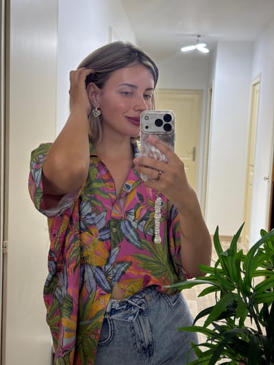 This is a mirror selfie of a young woman in Cluses, France. She is wearing a vibrant pink, green, and yellow floral print shirt and blue denim jeans. She is holding a clear phone case with a leopard print design in her right hand, which is capturing the reflection. The woman has blonde hair and is wearing subtle makeup, including lipstick. She has a diamond stud earring in her left ear and a ring on her right ring finger. Her left hand is resting on her hair, near her ear, and she has a slight smile on her face. A sparkly charm or bracelet is attached to her phone. In the background, visible in the mirror, are parts of an indoor setting, including a white door or closet, and a ceiling light fixture. A green leafy plant is also visible in the upper right portion of the image. The overall mood is casual and confident.