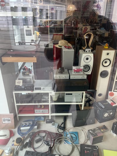 The image shows a shop window display in Wien, Austria (latitude: 48.202, longitude: 16.346). The display features a variety of audio equipment, including speakers, amplifiers, turntables, and cables. The audio equipment is arranged on a shelf and a table, with a red box, a white box, and a model car also on display. The image is taken from outside the shop, looking through the window. The window is reflective, showing a blurred image of the street outside. There is a blue and red cable coiled on the floor, along with other cables and a remote control on the shelf. The bottom right of the image shows a box labeled “Django Reinhardt,” and a yellow box labeled “Rocket” with a black label. The top right of the window shows a speaker with a black label reading “Tru Audio.”  A rubber duck is placed on top of the Tru Audio speaker, and the bottom right of the window displays a speaker with a “Model One” sticker. The display is illuminated by artificial light. The display is viewed through a glass storefront window with reflections. The image is slightly blurry.