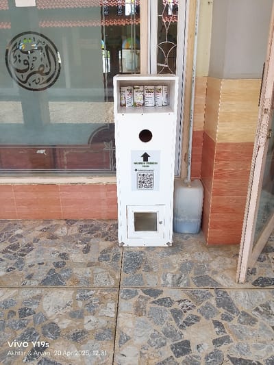 A white wooden box with a small window sits in front of a building in Kecamatan Haurgeulis, Indonesia (latitude: -6.46, longitude: 107.943). The box is attached to the wall and has a sign with Indonesian text and a QR code. Inside the box are six cans of a beverage, arranged in a row. The box is situated on a paved patio with a stone floor. Next to the box is a large white plastic jug. The building is constructed with light brown and orange tile. A large window with frosted glass is to the left of the box. The window has a decorative black and white pattern in the top right corner. There is a light brown area on the wall next to the box. The photo is taken from an angle that gives a clear view of the box and its contents.