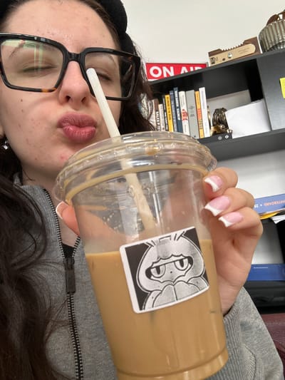 A slightly tilted, close-up portrait features a person drinking from a large, clear plastic cup with a white straw. The cup contains a light brown liquid, likely iced coffee, and bears a black and white sticker with an ornate, stylized design. The person's face is partially visible in the foreground, wearing large, dark-framed glasses, with eyes closed or looking downwards. Her lips are puckered around the straw. She has dark hair and is wearing a gray hooded garment; the zipper pull clearly displays the text "KARL." Her hand, holding the cup, features manicured nails with white French tips.
The background shows an indoor setting, likely a study or office space. A black shelf unit is visible, holding several books. Visible titles on the book spines include "REBEL," "VERNE HARNISH SCALING UP," "THE BUSINESS OF BELONGING," "BELONGING TO THE BRAND," "THE CULTURE CODE," and "SALES PITE." Chinese characters "中国" are also present on one book, and a binder titled "WHITEBOARD • 25" is visible further back. A partial red and white "ON AIR" sign can be seen on the left side of the shelf, along with a small wooden object marked "MM." The image captures a casual moment, possibly during a break from work or study in Antwerp, Belgium.