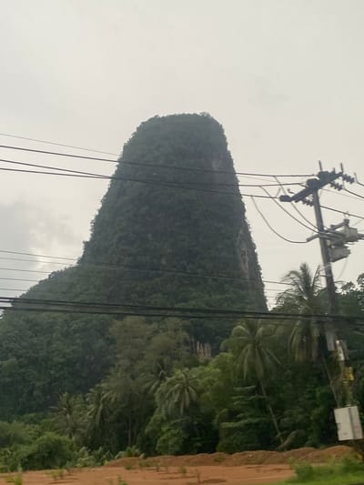 Captured from a ground-level perspective in Phang-nga, Thailand, the image features a tall, dome-shaped, heavily vegetated karst mountain rising prominently into a hazy, overcast sky. Power lines and utility poles intersect the foreground, obscuring portions of the mountain. Dense foliage, including palm trees, is visible on the mountainside and adjacent landscape. A dirt embankment is seen on the right side.