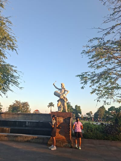 The image depicts an outdoor scene at the T B Kapal Public Park in Gianyar, Indonesia. The park features a prominent statue of a figure holding a drum and a drumstick, positioned atop a brick pedestal. In the foreground, two individuals are visible. One, a person wearing a dark t-shirt and shorts, stands facing left, looking at a smartphone. Another person, dressed in a purple t-shirt and black shorts, is seen from behind, facing right and appearing to be engaged in a light jog. The park environment includes paved pathways, tiered stone steps, lush green bushes, and trees, with a clear blue sky overhead. The sunlight casts long shadows, indicating it is either early morning or late afternoon.
