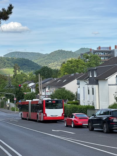 This media file captures a scene in Bonn, Germany, from a slightly elevated perspective. The primary subjects include a red and white public transport bus, a red sports car, and a black SUV parked along a road. In the background, residential buildings with dark tiled roofs are visible, interspersed with lush green trees. Beyond the buildings and trees, a series of rolling hills covered in dense forest dominate the landscape, stretching into the distance.

The image appears to be taken during daylight, with a partly cloudy sky. The lighting suggests it could be late afternoon or early evening, as the light is soft. The overall mood is serene and suggests a typical urban or suburban environment with natural elements.

Visible text includes markings on the vehicles and street signs. The bus has the number "612" and "602" displayed, along with a website address that appears to be "www.stadtwerke-bonn.de". The license plate of the red car reads "AH-GK-964". A speed limit sign indicates "50". Another sign features a "H" symbol, likely indicating a bus stop. A street sign reads "Oberstraße" and "Am Eichberg".

The orientation of the image is rotated 90 degrees clockwise. The scene depicts vehicles on a paved road, suggesting a street in a developed area, likely a residential neighborhood given the presence of houses and the natural backdrop of hills. No specific activity or event is clearly depicted; the vehicles are stationary, and the scene appears to be a moment captured in time.