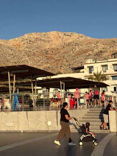 In Capaci, Italy, on a sunny day, a woman pushes a stroller containing a child along a paved walkway. Behind them, a group of children and adults, many wearing pink attire, are gathered on a raised area with a building in the background. A large, rocky mountain dominates the skyline. The scene appears to be a public space, possibly a promenade or plaza, adjacent to a coastal resort.