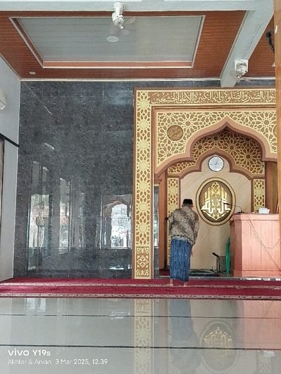 The image shows the interior of a mosque in Anjatan, Indonesia. The mosque has a grey marble wall on the left and a decorated niche on the right. The niche is decorated with intricate geometric patterns and Arabic calligraphy. A man in a brown and blue patterned shirt and a blue sarong stands in front of the niche, facing the wall. He is facing a prayer rug that lies on a tiled floor. There is a brown wooden podium to the right of the man with a book and some other items on top. On the wall inside the niche, there is a round plaque with Arabic calligraphy, a clock and a microphone. The ceiling is made of white and brown panels with a white ceiling fan in the middle of the room. The image is taken from a slightly low angle.