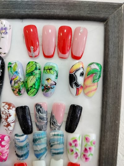 The image displays a collection of artificial nails, each adorned with various designs and colors. The nails are arranged in rows, with the top row featuring solid red and pink colors, some with subtle glitter accents. The second row showcases intricate hand-painted designs, including birds on branches, lush green foliage, and what appears to be a toucan. The third row presents a mix of patterns, from a floral design on a brown background to a black glitter nail, and abstract marbled patterns in shades of gray and pink. The bottom row continues with more abstract designs, including floral patterns on a white background and what seem to be marble patterns in blue and white. The nails are likely displayed for sale or as examples of nail art services. The background is a plain white surface, with a dark gray frame visible around the top and right edges of the image, suggesting the nails are presented within a display case or board. No people or specific activities are depicted, but the arrangement of the nails implies a retail or professional setting related to beauty and cosmetic services. The time of day, weather, and specific emotions are not discernible from the image. No visible text is present.