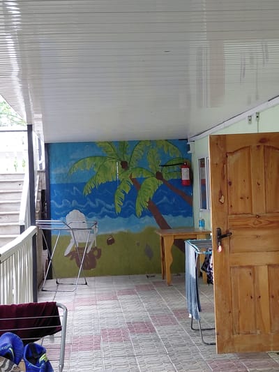The image depicts an outdoor covered patio area. The ceiling is made of white, horizontal panels. A mural on the back wall shows a tropical scene with palm trees, a blue ocean, and a green grassy area. In front of the mural, there is a white Adirondack-style chair and a portable drying rack with laundry. To the right, a wooden door with vertical panels and a dark handle is partially open. A red fire extinguisher is mounted on the wall near the door. The floor is tiled with a pattern of gray and pink squares. On the left side of the image, a white railing and a set of outdoor stairs are visible. Another drying rack is present in the foreground, holding a dark red piece of fabric and a pair of blue sandals. The location is identified as Ақши, Kazakhstan.