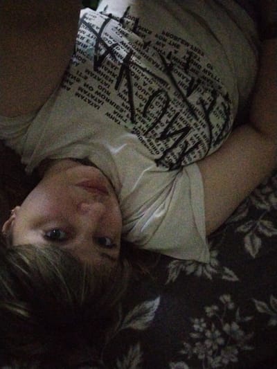 The image shows a person lying down on a floral-patterned surface. The person's head is tilted and they are looking towards the camera. They are wearing a light-colored t-shirt with a black graphic print, which appears to be text. The t-shirt print is partially obscured by the person's body and the way the t-shirt is creased. The person has dark hair, which is partially covering their face. The background is a dark, floral-patterned fabric, possibly a blanket or upholstery. The lighting is dim and appears to be coming from the side. There is no discernible information in the image that would indicate a specific location within the city of Yaroslavl, Russia.
