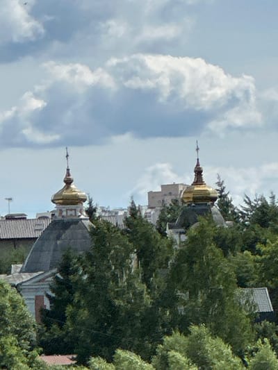 The image shows a view of the Church of the Sign of the Most Holy Theotokos in Dubrovitsy, Russia. The church features a distinctive golden dome topped with a cross, which is visible amongst a canopy of green trees. In the background, other buildings and structures are partially obscured by foliage and clouds. The sky is partly cloudy with dramatic white and grey formations against a light blue backdrop. The scene appears to be captured from a distance, likely from a higher vantage point.