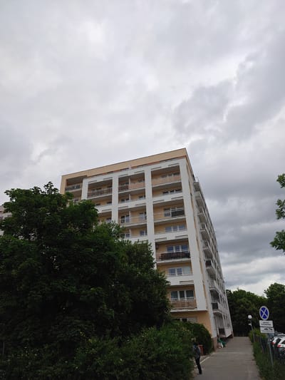 A low-angle, full shot captures a tall, modern apartment building in Poznan, Poland, under a heavy, overcast sky. The building is primarily white with tan accents and features numerous balconies with white railings. A large, dense tree with dark green foliage dominates the left side of the frame, partially obscuring the lower floors of the building. A paved walkway is visible in the lower right, leading away from the building. On the right side of the walkway, a traffic sign with Polish text indicates "Droga Pożarowa" (Fire Road) and "Zakaz parkowania" (No Parking). A person wearing a backpack stands on the walkway near the entrance to the building. Several parked cars are visible behind a fence on the far right. The overall scene suggests a typical urban residential environment.