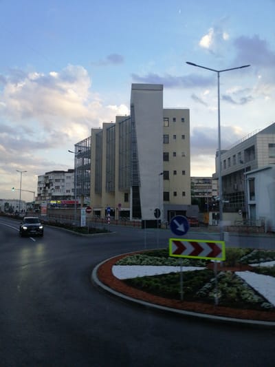 The image depicts a street scene in Fălticeni, Romania, likely during dusk given the lighting. A black car is driving on a wet asphalt road, heading towards the viewer. In the background, several modern buildings of varying heights are visible. One prominent building features a beige facade with a unique angular design and external metal grating. To its right, another building has a light-colored facade and numerous windows. Further in the distance, a lower, multi-story residential building with balconies can be seen. The sky is a soft blue with scattered clouds, and a crescent moon is visible. Streetlights and traffic signs are present, including a blue circular sign with a white arrow pointing left, and a horizontal rectangular sign with alternating red and white chevrons indicating a directional hazard. A roundabout with a landscaped island is also featured in the foreground. The overall impression is of an urban environment with contemporary architecture.