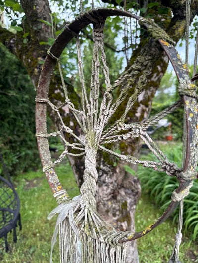 The image depicts an outdoor scene, likely a garden, in Užliekniai, Lithuania. The focal point is a rusted metal hoop, possibly part of an old wheel, decorated with macrame-style rope work. The hoop is suspended by thick ropes and has additional rope intricately woven across its diameter, creating a net-like pattern. Some sections of the rope show signs of wear and weathering.

The hoop is attached to a tree with a mossy trunk. The tree is leafy and appears to be in a natural setting, with green grass and foliage visible in the background. To the left, a black, woven chair or swing can be seen, partially out of focus. Further in the background, the environment suggests a rural or suburban outdoor space, with hints of other greenery and possibly structures. The overall impression is one of rustic decoration or a handcrafted garden feature.