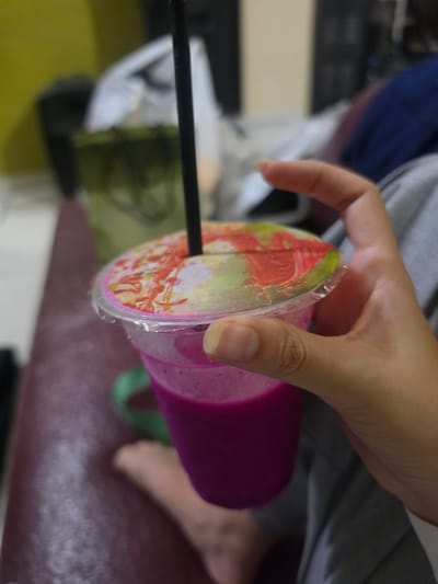 A close-up shot shows a person holding a clear plastic cup filled with a vibrant purple drink, likely a smoothie or juice, possibly made from dragon fruit. The cup has a colorful lid featuring a pattern of flowers and red and green abstract designs. A black straw extends from the lid, piercing the liquid. The person's hand, with visible fingernails, is positioned to hold the cup.
The background is blurred, but suggests an indoor setting with hints of furniture and possibly other objects. The lighting indicates it could be daytime, and the overall mood seems casual. The presence of what looks like a patterned fabric on the person's lower body, as well as the visible foot, adds to the impression of a relaxed, everyday moment. The image appears to be taken in Medan, Indonesia, though no specific landmarks are visible to confirm this. There is no visible text in the image.