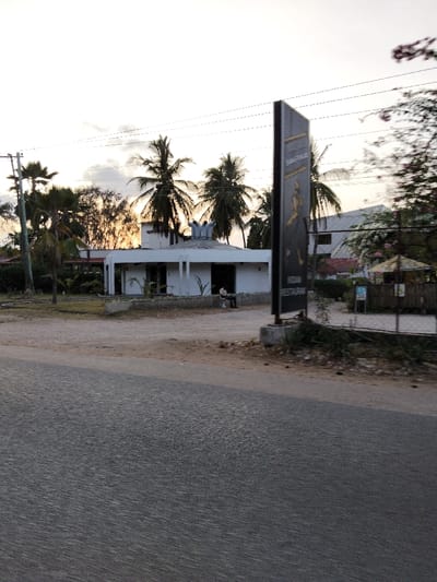 This image depicts a commercial roadside scene in Gombato Bongwe ward, Kenya, during dusk or dawn, characterized by a pale orange and white sky with scattered clouds. The foreground consists of a dark gray asphalt road surface. Beyond the road, a dirt and gravel area leads to the main subjects.

On the right, a large, dark vertical billboard is anchored by a concrete base. It displays "SHANGRILAS" and "INDIAN RESTAURANT" in white lettering, accompanied by a stylized dark figure. To the left of the billboard, a white, single-story building features a curved, modern facade and a circular roof with three cylindrical white structures protruding upwards. Two individuals are seated on a low concrete barrier or bench directly in front of this building. One person, dressed in dark clothing, faces another person in lighter attire.

Numerous tall palm trees are silhouetted against the sky, predominantly behind the white building and to the left. A utility pole with overhead power lines is visible on the far left. In the background, a larger, light-colored building is partially visible behind the billboard and the white structure. To the far right, a small, open-sided structure with a pointed, thatched-like roof stands, possibly a kiosk, with a smaller sign adjacent to it. Additional foliage, including some with pink blossoms, is present on the right edge.