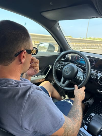 The image shows a person driving a Mercedes-Benz car. The perspective is from inside the car, looking over the shoulder of the driver. The driver is wearing a grey t-shirt and dark blue shorts. They have tattoos on their arm and are wearing sunglasses with gold accents on the side. The steering wheel and dashboard of the car are clearly visible. The car appears to be in motion, as suggested by the blurred scenery outside the window. The scenery outside consists of a road or elevated structure and some greenery, indicative of an outdoor setting. The time of day appears to be daytime, with bright sunlight. The overall mood is casual and relaxed. There is no visible text in the image. The location context is Capaci, Italy.
