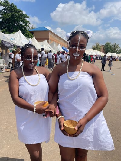 In Alagba, Nigeria, two young Black women are the primary subjects in this outdoor photograph, dressed in traditional white attire. They wear matching white beaded necklaces and bracelets, and their faces are adorned with white face paint, creating striking patterns around their eyes and mouths, and a central line down their noses and upper lips. Their hair is styled in braids, with white fabric wrapped around sections, giving a decorative, almost horn-like appearance.
The woman in the foreground is holding a hollowed-out gourd in her right hand, while her left hand rests on her lap. The woman behind her, slightly out of focus, holds a pink smartphone. Both women appear to be looking towards the camera with neutral or perhaps subtly smiling expressions.
The setting appears to be a daytime event, with a clear blue sky and scattered white clouds visible. The ground is paved, suggesting an outdoor gathering area or street. In the background, several white tents are set up, and a crowd of people, also mostly dressed in white or dark striped attire, are visible. A building with orange brick and a blue roof is present further in the background. The overall atmosphere seems festive, possibly a cultural celebration or ceremony.
There is no visible text in the image. The time of day appears to be mid-morning to afternoon, judging by the bright sunlight and shadows. The event is likely a community gathering, possibly a festival or ritual, given the traditional dress and face paint.