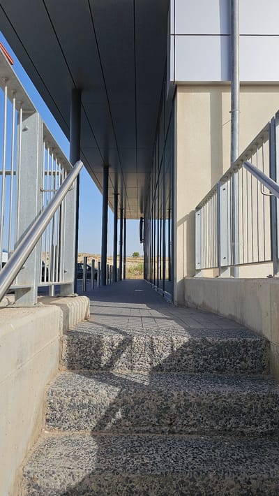 The image shows an outdoor scene with a modern building featuring a dark, sleek overhang supported by vertical columns. To the left, a metal railing with vertical bars and a curved handrail ascends a set of concrete steps. The steps themselves are textured with exposed aggregate. A walkway paved with square tiles stretches away from the steps, bordered by the building's glass facade on the right and more metal railings and bollards on the left. In the background, a dry, sparsely vegetated landscape with rolling hills under a clear blue sky can be seen. A few cars are parked in the distance. The sun casts sharp shadows from the railings and building structure onto the walkway and steps. The overall impression is one of a well-maintained public or commercial outdoor space, possibly near an entrance or pathway. The specific location in Cuarte de Huerva, Spain, is not visually identifiable from the image alone.