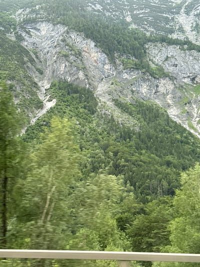 The image depicts a steep, rocky mountainside covered in dense green vegetation, primarily trees and shrubs, in Golling an der Salzach, Austria. The lower portion of the image is blurred due to motion, suggesting it was taken from a moving vehicle. A light-colored railing or barrier is visible at the bottom, further indicating a roadside perspective. The mountain exhibits significant rock formations and scree slopes interspersed with areas of forestation. The sky is overcast. There are no people or animals visible in the image.