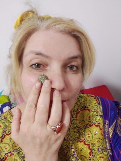 A medium close-up, head-and-shoulders shot shows a light-skinned person with blonde hair pulled up with a yellow accessory. The person is holding a textured, metallic-colored object to their right cheek with their left hand. The person's left ring finger wears a ring with a large, rectangular reddish-orange stone. The person is wearing a colorful, embroidered garment with gold, red, and purple paisley patterns. The background is a plain white wall and a red surface. There is no visible indication of the location in Al Mas'oudi, United Arab Emirates.