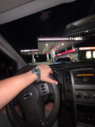 The image, taken from inside a vehicle at night, shows a person's arm and hand wearing a watch on the steering wheel of a car. The car appears to be at a LUKOIL gas station, the signage of which is visible through the windshield. The location is Momin Sbor, Bulgaria. The dashboard and interior of the car are also partially visible.