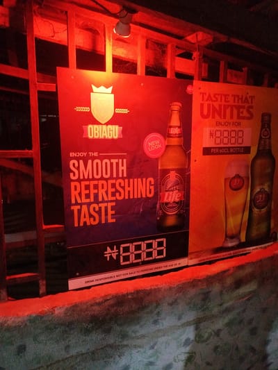 The image shows two advertising posters for alcoholic beverages, illuminated by red lighting. The poster on the left advertises "OBIAGU" and features text about "SMOOTH REFRESHING TASTE" along with a price of "#8888". A bottle of "Life" continental lager beer is also visible on this poster. The poster on the right advertises "TASTE THAT UNITES" and shows a price of "#8888 PER 60CL BOTTLE". This poster includes images of a glass of beer and two bottles, one of which is labeled "33". The posters are mounted on a wall or structure, and the surrounding environment appears to be an outdoor setting, possibly a bar or roadside establishment. The lighting casts a strong red hue over the entire scene. The text at the bottom of both posters states "DRINK RESPONSIBLY. NOT FOR SALE TO PERSONS UNDER THE AGE OF 18". The location of Akabuka, Nigeria is not visually discernible from the image itself, as no identifying landmarks or signs are present.