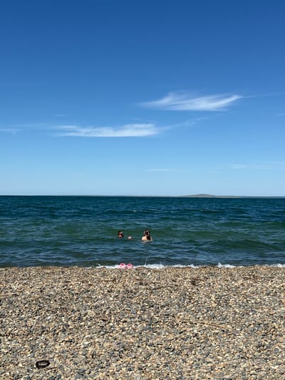 The image displays a serene coastal scene under a bright, clear sky, captured in what appears to be midday or early afternoon. The original image is rotated 90 degrees clockwise; when viewed correctly, it shows a horizontal composition divided into sky, sea, and pebble beach.

The upper portion of the image is dominated by a vast, bright blue sky featuring sparse, wispy white clouds. A clear horizon line separates the sky from the calm, deep blue-green waters of the sea. A faint outline of a distant landmass or hill is visible along the horizon.

In the sea, two individuals are seen partially submerged. One person, closer to the shore, is visible from the shoulders up, with dark hair, facing towards the beach. The other individual is further out in the water, less distinctly visible. The water itself shows gentle ripples, indicating calm conditions.

The lower part of the image features a textured pebble beach, extending from the water's edge. The pebbles vary in size and are a mix of grey, brown, and off-white tones. On the pebbles near the waterline, a pair of bright pink flip-flops are clearly visible. Further along the beach, a small, dark, circular object, possibly a hair tie or bracelet, rests on the stones. Scattered across the pebbles are also a few pieces of small natural debris.

The scene suggests a peaceful and sunny day at a beach, consistent with a leisure activity in a coastal area like Taman, Russia. No visible text is present in the media file.