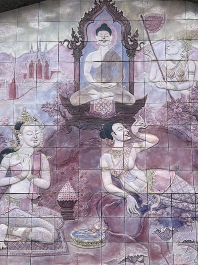 This is a photograph of a bas-relief tile mural depicting figures in what appears to be a traditional Thai or Southeast Asian artistic style. The mural is composed of numerous square tiles, creating a grid pattern over the entire artwork.

The central figure, positioned at the top, is a serene Buddha in meditation, seated on an ornate platform beneath an elaborate arch. The Buddha wears traditional monastic robes and has a halo or aura around his head.

To the right of the Buddha, another figure is depicted, also with a serene expression and adorned in ornate attire. This figure holds a long staff topped with a decorative fan-like object.

Below the Buddha, to the left, a kneeling figure with hands clasped in a devotional gesture faces forward. This figure wears a detailed headdress and traditional clothing.

To the right of the kneeling figure, and below the figure with the staff, another figure is shown reclining or seated in a dynamic pose, with one arm raised and head tilted back, displaying an expressive facial feature. This figure also wears elaborate traditional garments.

The background of the mural features stylized mountains, architectural structures resembling temples or pagodas, and floral motifs, including lotus flowers. The color palette is dominated by muted purples, pinks, blues, and grays, giving the artwork a somewhat aged or aged appearance.

The overall scene evokes a religious or mythological theme, common in Buddhist art and culture. The composition suggests a narrative or a depiction of celestial beings and significant figures. The location is specified as Tambon Ban Luang, Thailand, indicating this is likely an artwork found in a temple, public space, or cultural site in that region.