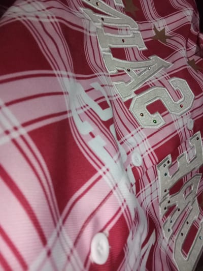 The image shows a close-up of a red and white plaid shirt. The shirt has large, white embroidered letters with sparkly embellishments, possibly rhinestones, in various colors like red, green, blue, and gold. Some of the letters are partially visible. There are also gold star shapes scattered on the fabric. Two white buttons are visible on the shirt. The fabric is creased, suggesting it might be worn or folded. The lighting is dim, creating shadows and highlighting the textures of the fabric and embroidery. The scene appears to be a still shot of the clothing item, with no people or specific setting evident beyond the fabric itself. There is no visible text apart from the embroidered letters, which are difficult to fully discern.