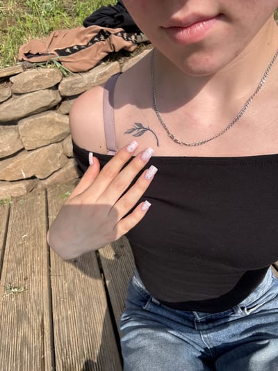 The image features a person laying down on a wooden surface outdoors. The person is wearing a black tank top and blue jeans. They have a silver chain necklace and a small tattoo of a leaf on their chest. Their fingernails are long and painted with a light pink and blue speckled design. The person's left hand is resting on their chest.
In the background, there is grass and some large rocks. To the left of the person, there are some pieces of clothing, including a tan jacket with a black logo. The scene appears to be outdoors during the daytime, as evidenced by the strong sunlight and shadows. The overall mood of the image is relaxed and casual. There is no visible text in the image.