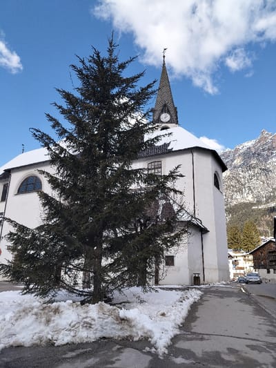 The image depicts a winter scene in Serdes, Italy, featuring a large evergreen tree in the foreground positioned slightly left of center. The tree, dark green with some brown tones, stands amidst irregular patches of dirty, partially melted snow covering the dark asphalt ground.

Behind the tree, a white-walled church building with a prominent steeple is visible. The church roof is dark and partially covered with snow. The steeple, featuring a dark conical roof, houses a circular clock face indicating approximately 11:20 or 11:30. A dark weather vane with an animal-like figure crowns the steeple's peak. Visible architectural details on the church include arched windows on the main edifice and rectangular windows on the tower section.

To the right of the church and in the distant background, snow-capped mountains rise under a bright blue sky dotted with scattered white clouds. A paved road or pathway runs along the right side of the image, showing remnants of snow. A few distant buildings with traditional architecture and some sparse evergreen trees are observable between the church and the mountains. No individuals are present in the scene.