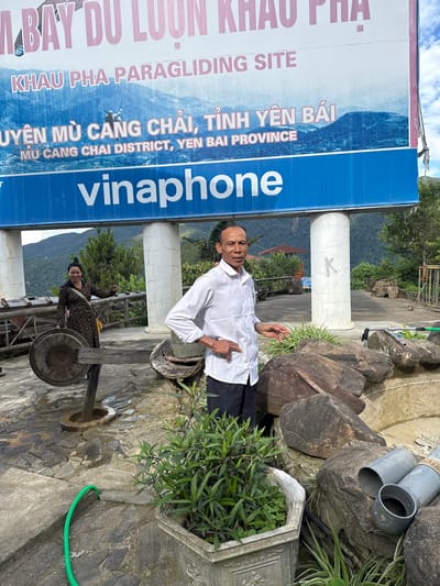 The image depicts the Khau Pha Paragliding Site in Yen Bai Province, Vietnam, located near the commune of Tu Le. A large billboard dominates the upper frame, displaying "KHAU PHA PARAGLIDING SITE" and "MÙ CANG CHAI DISTRICT, YEN BAI PROVINCE" in both Vietnamese and English text, alongside the "vinaphone" logo on a blue stripe. The billboard features a paraglider against a mountainous backdrop.
In the mid-ground, a dark-skinned adult male with short dark hair, wearing a light-colored checkered long-sleeved shirt and dark trousers, stands facing forward with his left hand on his hip and his right hand slightly raised. To his left, an adult female with dark hair styled in a bun, wearing a dark patterned dress and carrying a yellow bag, smiles towards the viewer while holding a stick or small object in her right hand. She stands next to a large, dark metal gong mounted on a wooden stand. The ground is paved with irregular grey stones, appearing wet in several areas. A green garden hose is visible on the ground in the lower-left foreground. A grey ornate planter containing green leafy plants is positioned in the lower foreground. Several large, dark rocks and two grey PVC pipes lie on the ground in the mid-right. Lush green vegetation, including trees and shrubs, is visible in the background, with distant mountains under a partially cloudy sky. The billboard structure is supported by two white cylindrical pillars, one of which has a black 'K' marked on it.