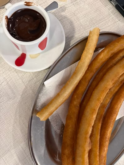 In Santa Ponça, Spain, a plate of churros sits next to a cup of hot chocolate. The churros are golden brown and appear to be fried dough, dusted with sugar. The hot chocolate is dark and rich, served in a white cup with red polka dots. The scene is set on a light-colored, textured surface, possibly a table covered with a tablecloth. The lighting suggests an indoor setting, perhaps a cafe or restaurant. No people are visible in the image, but the presence of the food implies it is being served or consumed.