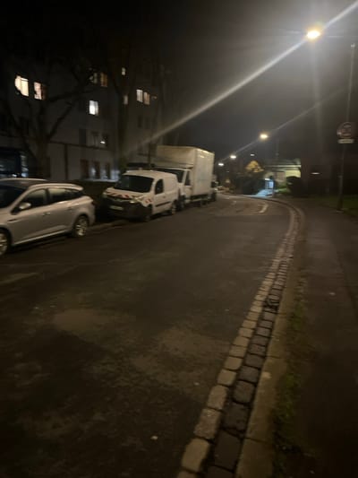 A nighttime urban street scene. Cars and vans are parked along the left side of the road, with building facades visible behind them. Streetlights illuminate the scene, casting long shadows. The road surface appears wet or damp, reflecting the light. A curb made of rectangular stones lines the edge of the pavement. A speed limit sign is visible at the top. The image is slightly rotated, giving a tilted perspective.