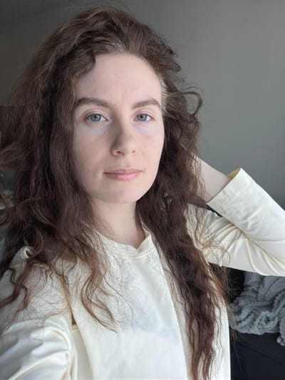 The scene is a close-up indoor portrait of a female subject, located in Yanino-1, Russia. The individual, appearing to be a young adult, has long, wavy dark brown hair with noticeable graying strands near the temples and part. Her complexion is pale, featuring light blue eyes and light-colored lips; no prominent makeup is apparent. She wears a light cream-colored, long-sleeved top with a visible seam detail near the collar. Her left arm is bent, with the hand positioned behind her head or neck, partially visible on the right side of the frame. She faces forward, looking directly into the camera with a neutral to slightly soft expression. The background consists primarily of a plain light gray wall. To the lower right, behind the subject's arm, a dark object, possibly furniture, is partially visible, draped with a textured gray fabric.