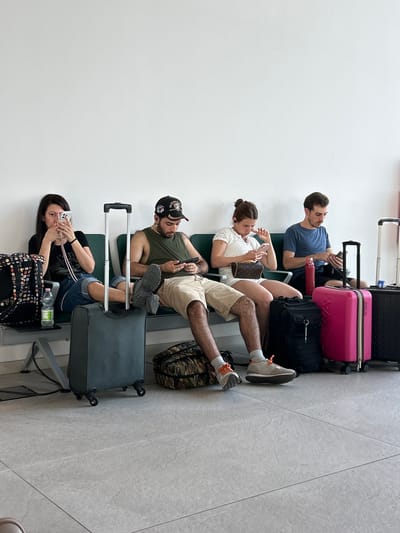 Four people are seated in an airport terminal, awaiting their flight. The scene is brightly lit, suggesting it is daytime. The floor is tiled with large, light grey tiles. The people are sitting on dark green seats, with their luggage arranged around them.

From top to bottom:
A man in a blue t-shirt is looking at his phone, with a pink suitcase and black bag to his right.
A woman in a white top is also looking at her phone, holding a Louis Vuitton handbag.
A man wearing a green tank top and a baseball cap is looking at his phone, with a camouflage duffel bag to his right.
A woman in a black t-shirt is looking at her phone, with a patterned bag and a water bottle beside her.

The luggage includes a pink suitcase, a black suitcase, a duffel bag, and a patterned bag. The overall mood appears to be relaxed, with each person occupied with their phone, a common sight in airport waiting areas. The presence of luggage indicates travel, and the bright lighting suggests daytime operations within the airport. There is no visible text in the image. This scene is set in Lamezia Terme, Italy.