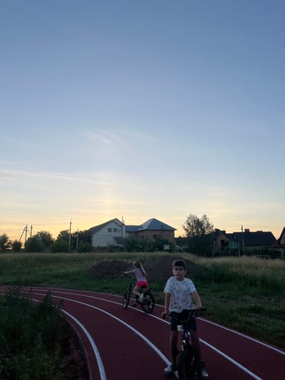 The image depicts two children on bicycles on a running track during what appears to be sunset. The boy in the foreground is facing the camera, wearing a patterned t-shirt and dark shorts, and is on a bicycle. A girl on a bicycle is in the background, riding away from the camera. The running track is red with white lane markers. Behind the track, there is a grassy field with scattered trees and several houses visible in the distance. The sky is a gradient of blue and orange, indicating the time of day. The location is identified as Koroviia, Ukraine.
