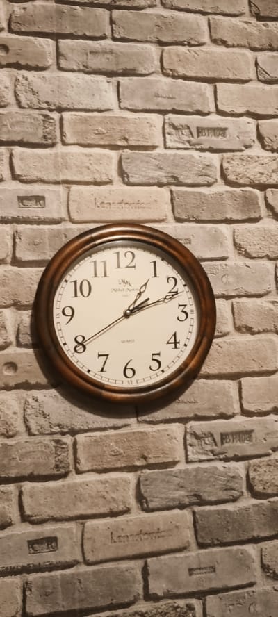 A round, dark wooden clock is mounted on a brick wall. The clock has a white face with black numbers and hands. The hour hand points to 1:40, and the minute hand points to 8. The wall is made of rough, textured bricks in shades of gray and brown. Some of the bricks have inscriptions or stamps on them. The lighting suggests an indoor environment, possibly a home or office. The image does not depict any people or specific actions. No information is available to determine the location in Tver, Russia.