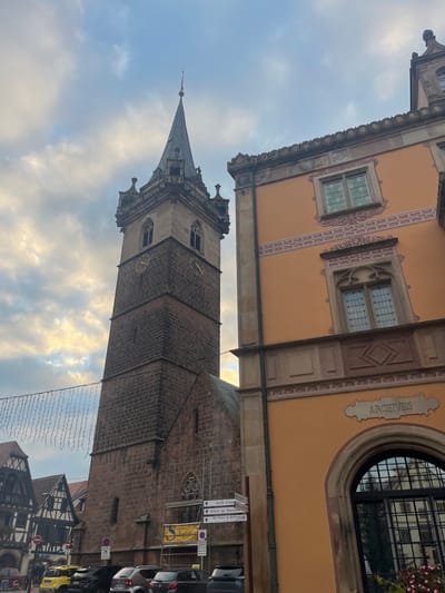 The image depicts a street scene in Obernai, France, focusing on two prominent architectural structures under an overcast sky with patches of bright light.

On the left, a tall, square, multi-tiered stone tower, identified as the Kappelturm, rises into the sky. Its lower sections are constructed from rough-hewn reddish-brown stone blocks. Higher up, the tower transitions to lighter stone, featuring four clock faces with golden numerals and hands, two of which are partially visible. Above the clock level, ornate gothic windows are set into the facade, topped by a steeply pitched, dark-colored spire. Scaffolding is partially visible at the base of the tower.

To the right, a multi-story building with a warm orange facade dominates the mid-ground. This structure, the former Halle aux Blés now housing the municipal archives, displays contrasting light-colored architectural detailing around its windows and corners. The upper level features rectangular windows with decorative surrounds. A horizontal decorative band with a repeating scroll pattern runs below these windows. The lower level includes a large arched window with black grilles, reflecting the surroundings. Above this arch, the word "ARCHIVES" is embossed within a decorative panel.

In the foreground at street level, several vehicles are parked. A multi-directional white street signpost points to local destinations such as "Salle d'expo" and "Office de Tourisme." Overhead, string lights are suspended horizontally across the street, suggesting urban decoration. Further left in the background, traditional half-timbered buildings characteristic of the Alsace region are partially visible. No individuals are clearly identifiable in the scene.