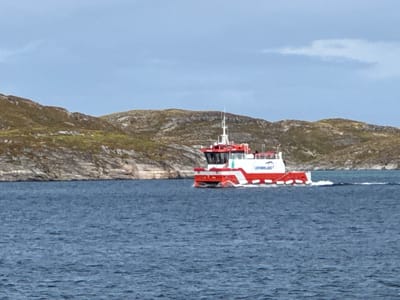 A white and red boat, named "LOFOTBLAST," travels on a choppy blue ocean. The boat has a prominent white structure at the front with windows and a red roof. The back of the boat is predominantly red with white accents. The boat is leaving a white wake behind it. In the background, a rocky, sparsely vegetated hillside with brown and green tones rises from the water. The sky above is overcast and a light blue. The image appears to be taken from another boat or the shore, looking towards the moving vessel. The scene suggests a maritime setting, possibly in a coastal area of Norway like Lovund, known for its rugged natural beauty and maritime activities.