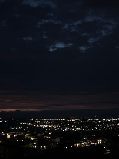 This is a nighttime aerial view of a city at dusk. The sky is a deep, dark blue with scattered clouds, and a faint red glow from the setting sun is visible on the horizon. Below, the city lights twinkle, creating a sprawling tapestry of white and yellow dots. In the foreground, houses with illuminated windows are visible, suggesting a residential area. In the distance, mountains form a dark silhouette against the dim light. The image appears to be taken from a high vantage point, offering a panoramic perspective of the urban landscape. The overall impression is one of a peaceful evening scene, with the city coming alive as night falls. The location is the city of Markovo, Bulgaria.