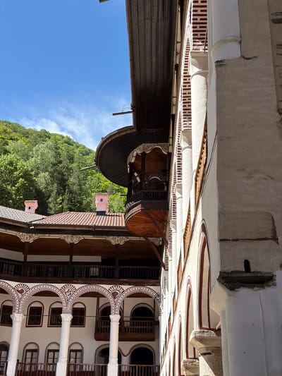 The image shows the courtyard of the Rila Monastery in Rilski manastir, Bulgaria, with the sky and a forested mountain visible in the background. The architectural details include red and white striped arches and columns, wooden balconies with decorative carvings, and a well with a prominent wooden structure. The roofs are tiled in red. The sky is clear blue with a few clouds.