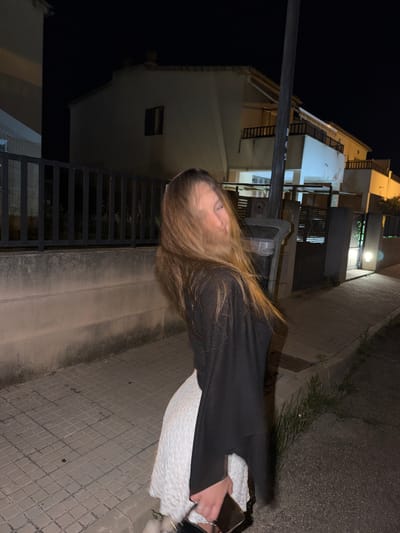 A woman stands on a tiled sidewalk in Platja D'alcúdia, Spain, at night. She wears a black top and a white skirt. She is holding a phone in her right hand. Buildings line the street, illuminated by streetlights. A black metal fence is visible in the foreground.