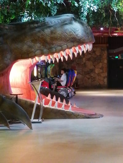 A person is operating a ride inside a large dinosaur head structure, which is situated indoors in Hong Gai Ward, Vietnam. The dinosaur head has large, prominent teeth. The setting appears to be a recreational or amusement area. The person is seated in a ride with wheels that has flashing lights and colored seats, and the ride is positioned inside the open mouth of the dinosaur. The environment is indoors, with interior lighting and building details visible in the background, including a stone-like wall and a doorway. A structure similar to a drink stand can be seen on the left inside of the mouth.