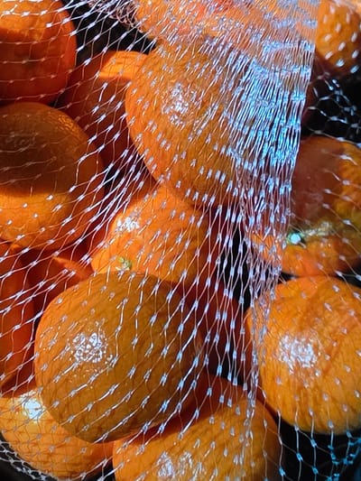 A close-up, over-the-shoulder shot captures a bag of clementines, packed tightly and illuminated by an orange-toned light. The mesh bag, a common packaging for such produce, is a prominent feature, with its white netting creating a diagonal pattern across the frame. Through the mesh, the vibrant orange of the clementines is visible, some appearing smooth and others with a slightly textured peel. The lighting creates a warm, inviting glow, highlighting the round shapes of the fruit and casting subtle reflections on their surfaces. The background is dark, further emphasizing the luminous quality of the clementines and their mesh container. No people are visible in the image. The scene does not contain any identifiable landmarks or elements that would suggest a specific location in Voluntari, Romania.