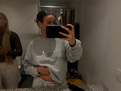 The image shows two women in a bathroom in Skanderborg, Denmark. The woman on the left has long blonde hair and is wearing a brown long-sleeved shirt and grey sweatpants. She is standing in front of the bathroom mirror, facing the camera. The woman on the right is holding a black smartphone in her right hand and taking a selfie. She is wearing a grey sweatshirt with a white graphic design, a white bow in her hair, and grey sweatpants. The background shows the bathroom in the background. A white electrical outlet is visible on the wall to the right side of the image. The bathroom is tiled and has a light brown door with white vertical blinds. There are also some clothes hanging on a rack in the background.