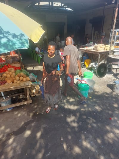 In Picha Ya Ndege, Tanzania, a street market scene unfolds under bright sunlight. Two individuals are the primary subjects. A man, wearing a grey t-shirt and red shorts, is lying on the ground, possibly resting. Beside him, a woman, dressed in a black t-shirt and a long grey skirt, stands and looks towards the camera. She is wearing flip-flops.

Scattered around them are various objects. Several large plastic containers, some green and some silver, are visible, along with what appears to be a metal basin. A yellow gas cylinder is also present. In the foreground, a vibrant umbrella provides shade, displaying colors of yellow and light blue. Behind the woman, a market stall is laden with produce, including coconuts, tomatoes, and what look like onions and garlic. Crates and wooden structures form the market's framework.

The ground is a mix of dirt and concrete, with scattered leaves and debris. Shadows from overhead structures and possibly trees are cast across the scene, suggesting a time of day when the sun is high. The overall impression is of a busy, outdoor market environment. The woman's direct gaze suggests she is aware of being photographed. No legible text is visible in the provided image.