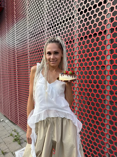 A woman with blonde braided hair stands in front of a red honeycomb-patterned wall, holding a small cake topped with strawberries and blueberries. She is wearing a white ruffled top and beige pants. The background appears to be an outdoor urban setting, with a cobblestone path and some greenery visible on the ground. The image was taken in Есбол ауылдық округі, Kazakhstan.