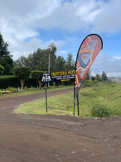 The image displays an outdoor scene in Makuyu, Kenya, featuring a dirt road alongside a green, grassy area. Two prominent signs are visible, advertising local businesses.

On the left, a tall, teardrop-shaped feather banner showcases a vibrant, patterned design, suggestive of traditional artwork. The text "GuChokia Rui MuKARO" is repeatedly printed on its surface. Next to this, a black rectangular sign with bright yellow lettering announces "TROTTERS HOT." Below this, white text lists the services provided: "ACCOMMODATION - CONFERENCE - SWI[MMING]", followed by "GARDENS - BAR & RESTAURANT." A stylized logo, possibly depicting a "TR" or two figures, is affixed to the left of the Trotters Hot sign.

The background features lush green trees and foliage, with power lines and utility poles stretching across the upper portion of the scene. The sky is blue with scattered white clouds, indicating it is daytime. In the distance, a road and a road sign are visible on a slight incline. The dirt road, with tire tracks, occupies the right side of the frame, suggesting a path leading to the advertised establishments. The overall setting appears to be a rural or semi-rural entrance to a commercial property.