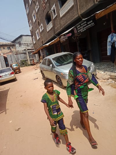 A mother and child walk hand-in-hand down an unpaved, reddish-brown dirt road in Awada, Nigeria. The adult female, with braided hair, wears a green, blue, and red patterned traditional outfit and carries an infant wrapped in matching fabric on her back. She wears black flip-flops. The male child, wearing a matching patterned green and blue outfit with red and black sneakers, walks to her left, holding her left hand. A silver Toyota Corolla sedan is parked facing away from the camera on the right, partially obstructing storefronts, one identified as "Glamplay Lounge" with services like "BARBING, DREAD LOCK." A multi-story brown building with white window frames and balconies lines the right side of the street. Further down the road on the left, a white sedan and a yellow three-wheeled vehicle are visible. Overhead utility wires traverse the clear sky. Several indistinct figures are present in the background.